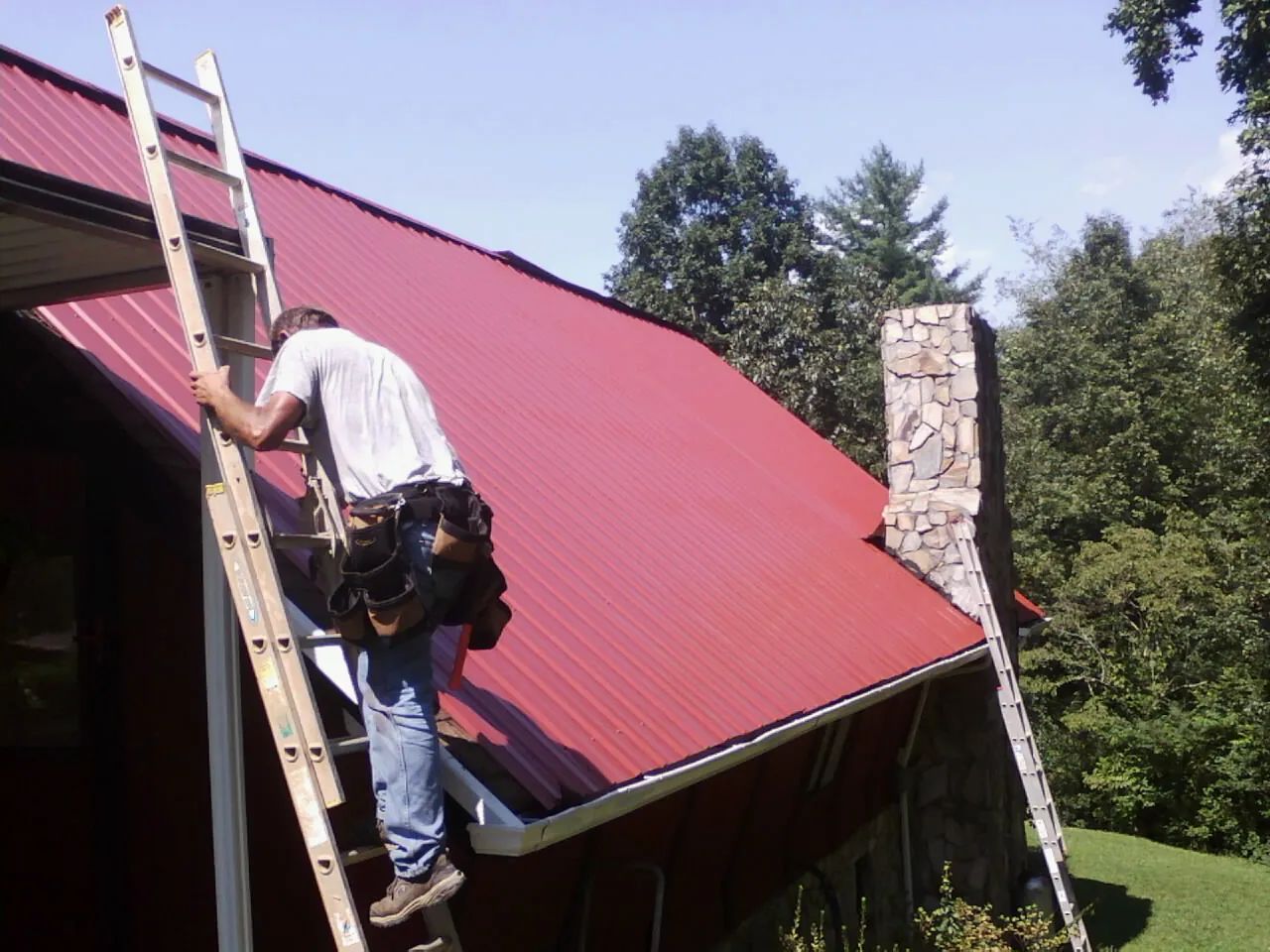 Roofer on a ladder beside a red metal roof and stone chimney — Soffit & Fascia Repair by Roofing Brothers in Pleasant Hall